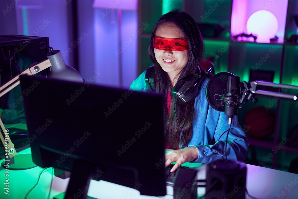 Sticker Young chinese woman streamer playing video game using virtual reality glasses at gaming room