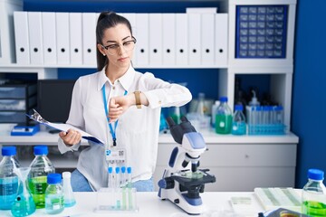 Young brunette woman working at scientist laboratory checking the time on wrist watch, relaxed and confident
