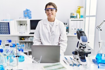 Young hispanic woman wearing scientist uniform working at laboratory looking positive and happy...