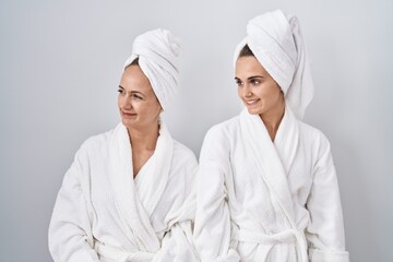 Middle age woman and daughter wearing white bathrobe and towel looking away to side with smile on face, natural expression. laughing confident.