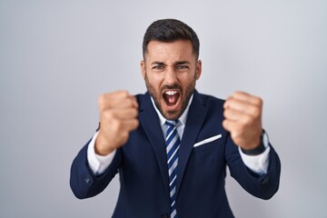 Handsome hispanic man wearing suit and tie angry and mad raising fists frustrated and furious while shouting with anger. rage and aggressive concept.