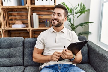 Handsome hispanic man holding clipboard working at psychology clinic looking away to side with smile on face, natural expression. laughing confident.