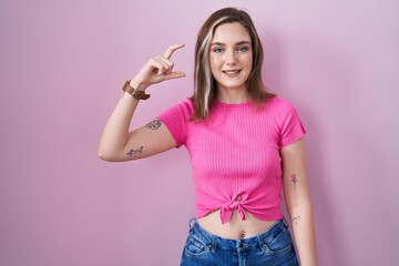 Blonde caucasian woman standing over pink background smiling and confident gesturing with hand doing small size sign with fingers looking and the camera. measure concept.