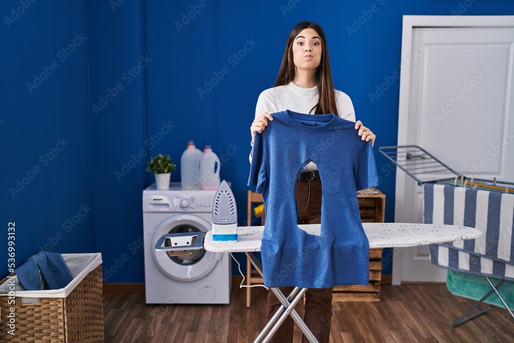 Poster Young brunette woman ironing holding burned iron shirt at laundry room puffing cheeks with funny face. mouth inflated with air, catching air.