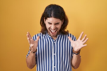 Hispanic man with long hair standing over yellow background celebrating mad and crazy for success with arms raised and closed eyes screaming excited. winner concept