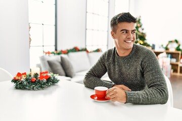 Young hispanic man drinking coffee sitting by christmas tree at home