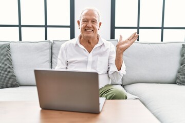 Senior man using laptop at home sitting on the sofa smiling cheerful presenting and pointing with palm of hand looking at the camera.