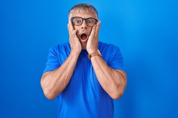 Hispanic man with grey hair standing over blue background afraid and shocked, surprise and amazed expression with hands on face