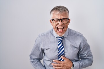 Hispanic business man with grey hair wearing glasses smiling and laughing hard out loud because funny crazy joke with hands on body.