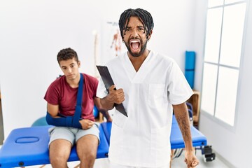 Young hispanic man working at pain recovery clinic with a man with broken arm angry and mad screaming frustrated and furious, shouting with anger. rage and aggressive concept.