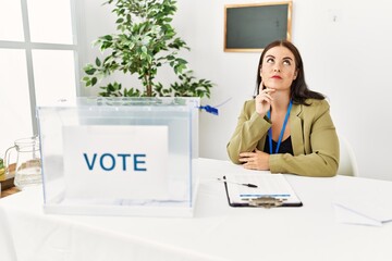 Young brunette woman sitting at election table with voting ballot with hand on chin thinking about...