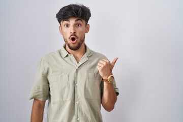 Arab man with beard standing over white background surprised pointing with hand finger to the side, open mouth amazed expression.