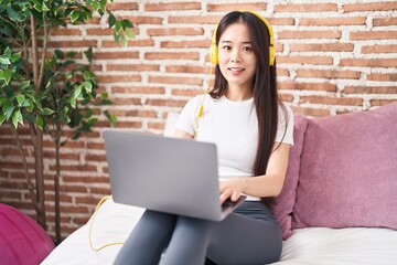 Young chinese woman using laptop and headphones sitting on bed at bedroom