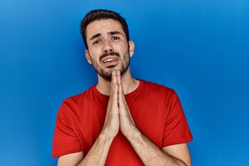 Young hispanic man with beard wearing red t shirt over blue background begging and praying with...