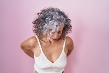 Middle age woman with grey hair standing over pink background suffering of backache, touching back...