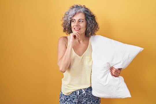 Middle age woman with grey hair wearing pijama hugging pillow with hand on chin thinking about question, pensive expression. smiling and thoughtful face. doubt concept.