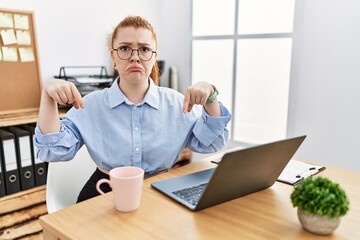 Young redhead woman working at the office using computer laptop pointing down looking sad and...
