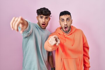 Young hispanic gay couple standing over pink background pointing with finger surprised ahead, open mouth amazed expression, something on the front