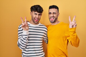 Young hispanic gay couple standing over yellow background smiling with happy face winking at the camera doing victory sign with fingers. number two.