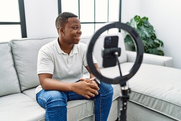 Young african man recording vlog tutorial with smartphone at home looking away to side with smile...