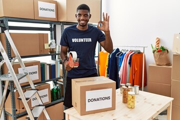 Young african american volunteer man packing donations box for charity smiling positive doing ok sign with hand and fingers. successful expression.