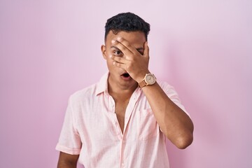 Young hispanic man standing over pink background peeking in shock covering face and eyes with hand,...