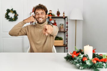 Young handsome man with beard sitting on the table by christmas decoration smiling doing talking on the telephone gesture and pointing to you. call me.