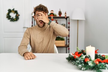 Young handsome man with beard sitting on the table by christmas decoration peeking in shock...