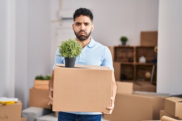 Hispanic man with beard moving to a new home holding box puffing cheeks with funny face. mouth...