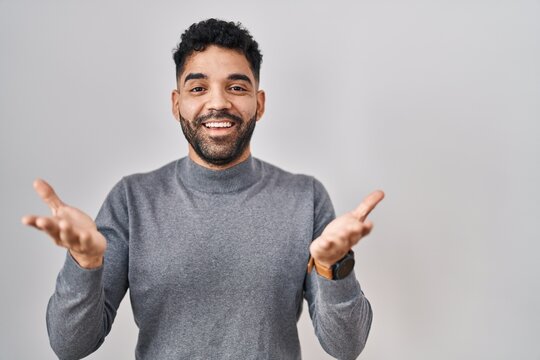 Hispanic man with beard standing over white background smiling cheerful with open arms as friendly welcome, positive and confident greetings