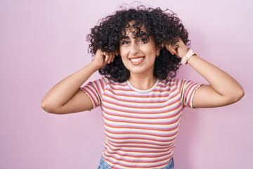 Young middle east woman standing over pink background smiling pulling ears with fingers, funny...
