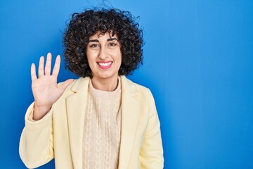Fototapeta na wymiar Young brunette woman with curly hair standing over blue background showing and pointing up with fingers number five while smiling confident and happy.