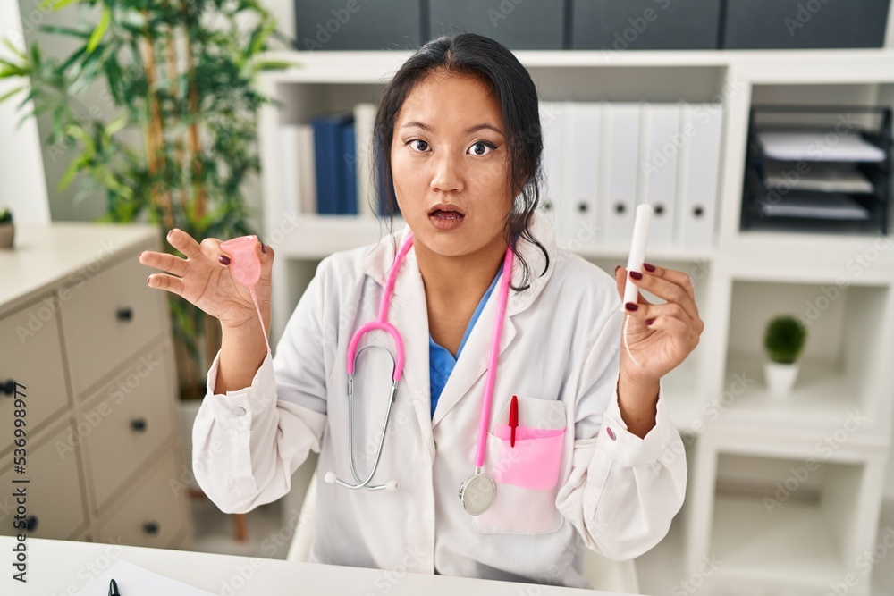 Wall mural young asian doctor woman holding menstrual cup and tampon in shock face, looking skeptical and sarca