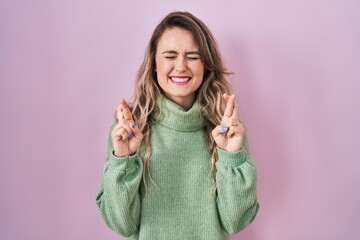 Young caucasian woman standing over pink background gesturing finger crossed smiling with hope and eyes closed. luck and superstitious concept.
