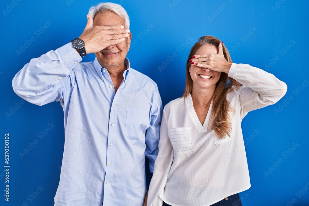 Canvas Prints Middle age hispanic couple standing over blue background smiling and laughing with hand on face covering eyes for surprise. blind concept.