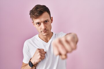 Caucasian man standing over pink background punching fist to fight, aggressive and angry attack,...