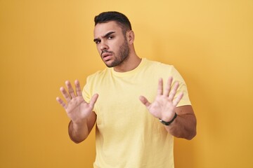 Young hispanic man standing over yellow background moving away hands palms showing refusal and...