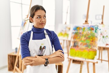 Young brunette woman at art studio looking to the side with arms crossed convinced and confident
