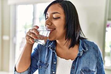 Hispanic brunette woman drinking a glass of water at the kitchen