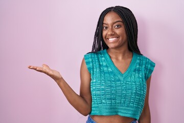 Young african american with braids standing over pink background smiling cheerful presenting and pointing with palm of hand looking at the camera.
