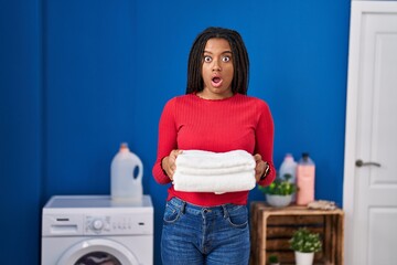Young african american with braids holding clean laundry in shock face, looking skeptical and sarcastic, surprised with open mouth