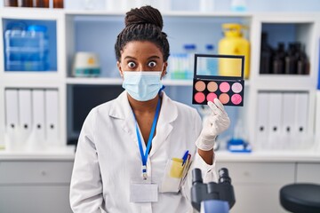 Young african american with braids working at scientist laboratory doing make up scared and amazed with open mouth for surprise, disbelief face