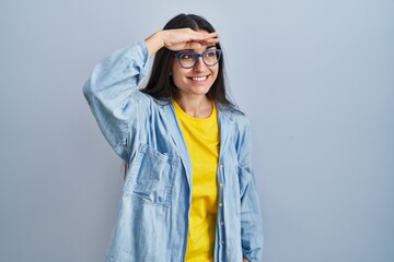 Young hispanic woman standing over blue background very happy and smiling looking far away with hand over head. searching concept.