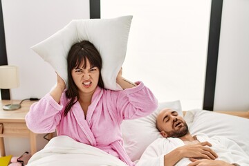 Hispanic woman angry with pillow on his ears while man snoring on the bed.