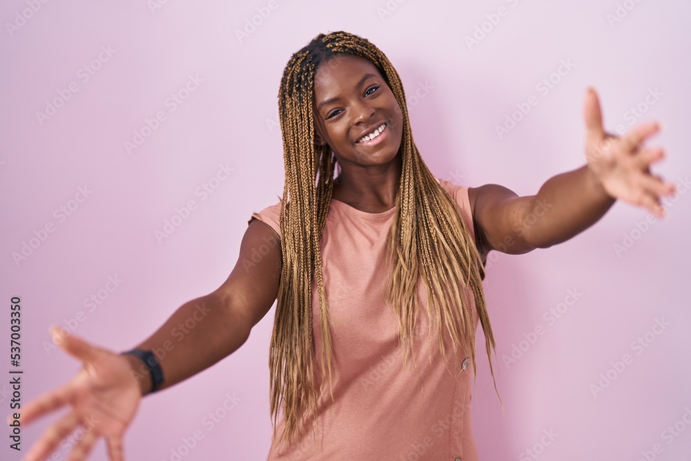 Sticker African american woman with braided hair standing over pink background looking at the camera smiling with open arms for hug. cheerful expression embracing happiness.