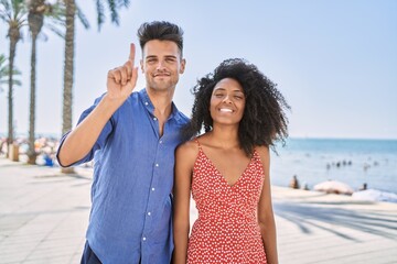 Young interracial couple outdoors on a sunny day showing and pointing up with finger number one while smiling confident and happy.