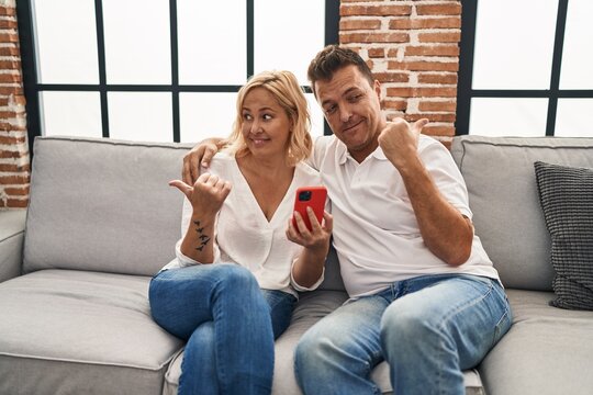 Middle age man and woman using smartphone sitting on the sofa pointing thumb up to the side smiling happy with open mouth