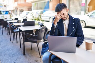 Young hispanic man using laptop with serious expression at coffee shop terrace