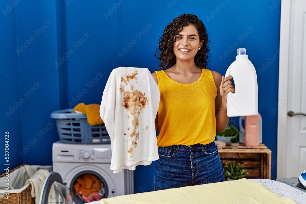 Wall mural young latin woman holding dirty t shirt and detergent bottle at laundry room - Wall murals