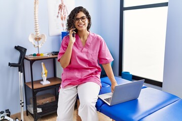 Young latin woman wearing physiotherapist uniform talking on the smartphone and using laptop at...
